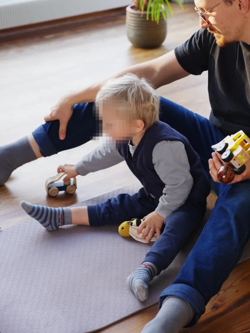 Andri seated behind an older child during a sitting exercise with a wooden toy